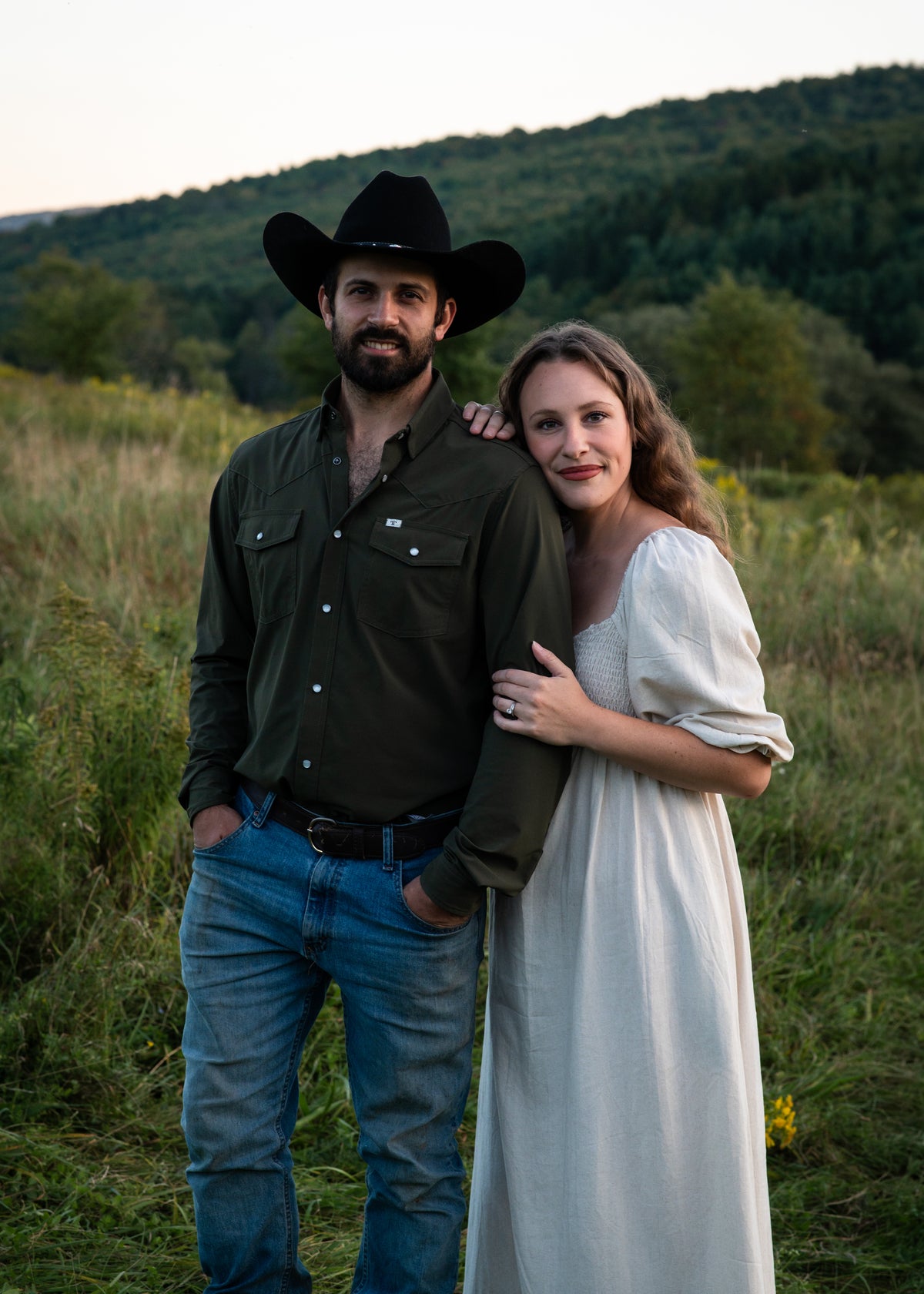 A couple standing outdoors in a grassy field with hills in the background. The man wears a black cowboy hat, dark green button-up shirt, and blue jeans, while the woman wears a long cream dress.